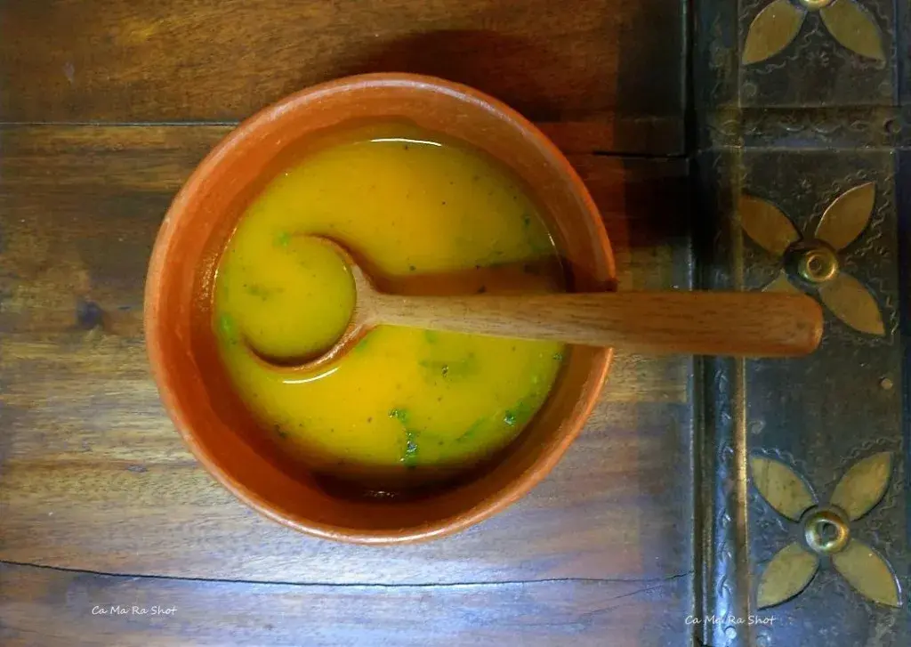A small terracotta bowl of golden rasam with a wooden ladle